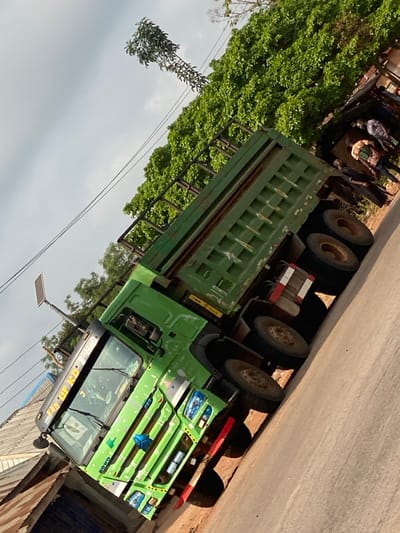 The image captures a roadside scene in Okura, Nigeria, centered on a large, green HOWO brand dump truck. The truck is positioned on an unpaved, light brown dirt or gravel road, facing toward the left side of the frame with its front angled upwards. The truck features a multi-axle configuration, with at least three visible rear axles fitted with dual wheels. Markings on the sun visor read "RNG UP KD RHO," and reflective red and white striped tape is present on the rear chassis.
To the immediate left of the truck's cabin, part of a building with a corrugated metal roof is visible. A solar-powered streetlight on a pole stands nearby. The background behind the truck is dominated by dense green foliage, likely trees or bushes. A taller, slender tree with sparse leaves extends above the main foliage line against a hazy, light grey sky.
On the right side of the image, near the rear of the truck, a group of approximately three to four individuals is visible standing on the dirt shoulder beneath the trees. They appear to be engaged in a stationary interaction or observation. One individual is identifiable by a patterned shirt. The overall lighting suggests daylight conditions.