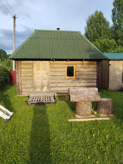 The image depicts a small, rustic wooden cabin with a green corrugated metal roof in Novaya Prismara, Russia. The cabin is constructed from logs, and it has a wooden door with two panels and a small rectangular window with a wooden frame. In front of the cabin, there is a wooden pallet serving as a step. To the right of the cabin, there is a bench made from a log supported by two tree stumps. The ground around the cabin is covered in green grass. In the background, there are trees and a telephone pole with wires. To the right of the main cabin, a portion of another structure with a similar green roof is visible. A portion of a silver ladder is visible in the lower left corner of the image. The sky above is partly cloudy.