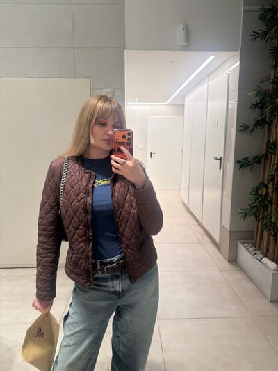 A woman takes a selfie in a bathroom mirror. She is wearing a brown quilted jacket, a dark blue shirt, and light blue jeans with a belt. She holds an orange phone with a patterned case and a tan bag with a teddy bear graphic on it. The bathroom features white tile floors and walls, with a green plant visible near the ceiling. There are also multiple white doors with black handles visible in the mirror.