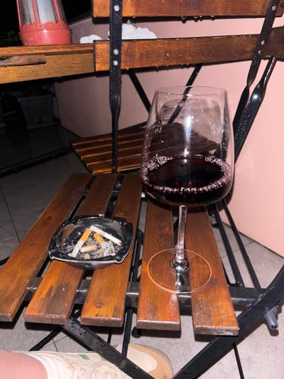 The image depicts a nighttime outdoor scene, viewed from a low angle, focusing on a wet surface with two primary objects. A clear, stemmed wine glass, approximately half-full of dark red wine with visible bubbles around the liquid's surface, stands on a wet, slatted wooden surface. To its left, a black, square ashtray contains four cigarette butts and a quantity of ash and debris. Both the wooden slats and an underlying dark metal frame exhibit numerous water droplets, indicating recent precipitation or heavy condensation.
In the upper left background, another higher, wet wooden surface is partially visible, holding a red, cylindrical object resembling a lantern or small heater, along with a small patch of light-colored, possibly frozen, material. A blurry, light pink wall or building facade forms the main background, with darker, indistinct shapes below the upper wooden surface. In the lower left corner, a partial view of a human leg and foot is visible, wearing light-colored footwear with a textured sole and what appears to be a light-colored sock or garment hem.
The image contains no discernible landmarks or specific visual cues to identify the location as the city of Nesso, Italy.