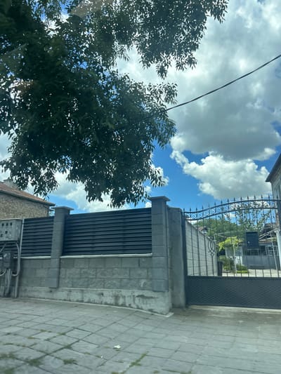 The image shows a view looking up towards a sky with white clouds and some dark green tree branches partially obscuring the view. In the foreground, a grey stone wall is topped with a dark horizontal slatted fence. To the right, there is a decorative wrought iron gate. Beyond the gate, there are glimpses of trees and buildings. The ground in the foreground is paved with square grey tiles. A grey electrical box is attached to the left side of the stone wall. The overall impression is an outdoor scene, possibly on a street or in front of a property, with elements of both modern and traditional design. The image is taken from a slightly low angle, looking upwards.