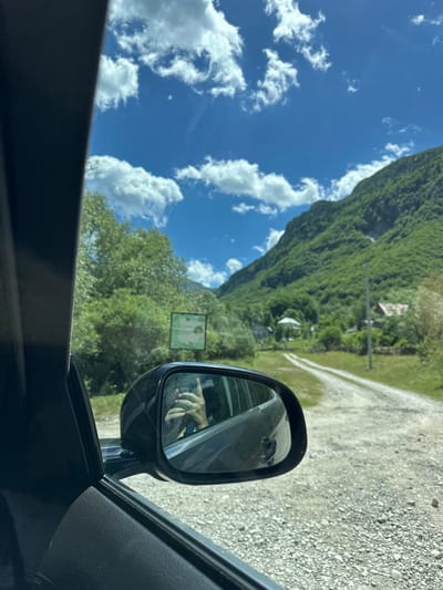 A side-view mirror of a car is visible on the left, reflecting a person holding a phone and taking a selfie. The mirror is angled to show a dirt road winding through a green, mountainous landscape. Trees and some houses are scattered along the road, and a sign is partially visible in the distance. The sky is blue with scattered white clouds. The scene is likely in or around Gusinje, Montenegro, given the characteristic mountainous terrain.