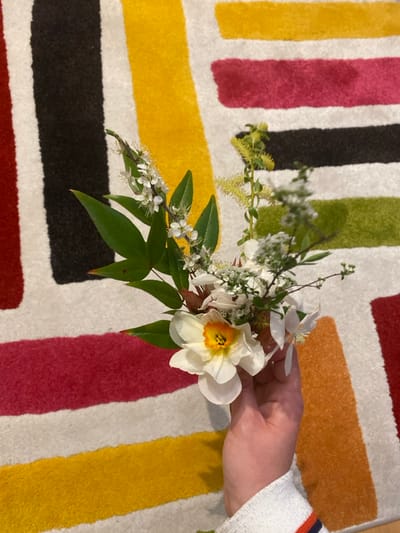 A person's left hand, observed from an elevated perspective, holds a small, mixed floral bouquet against a patterned rug. The hand exhibits light skin pigmentation and the cuff of a white long-sleeved garment with red/orange and blue stripes is visible at the wrist. The bouquet consists of one prominent white flower with a yellow-orange center, resembling a daffodil, interspersed with multiple sprigs of small white blossoms, various green leaves, and subtle reddish-brown foliage. The background features a plush rug characterized by broad, irregular vertical and horizontal stripes in colors including black, yellow, fuchsia, olive green, and orange, set against a white ground. The scene is situated indoors, within Boulogne-Billancourt, France.