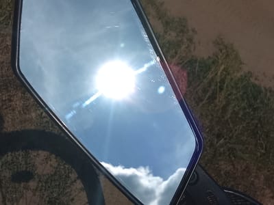 The image is a close-up, slightly angled shot of a motorcycle rearview mirror reflecting a bright, sunny sky with clouds and lens flares. The mirror is housed in a dark casing, and a portion of the motorcycle's handlebars and a dark shadow that appears to be a wheel are visible in the foreground. The background, outside of the mirror's reflection, shows dry grass and some vegetation. The setting is likely outdoors, given the natural light and vegetation. No specific location within Патоцкі сельскі Савет, Belarus, can be determined from the image alone.