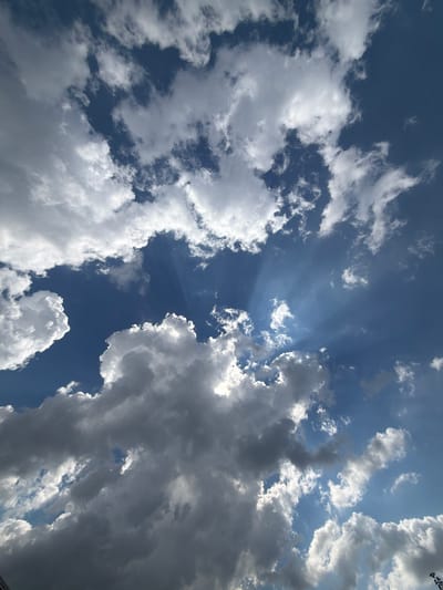 A low-angle, full shot captures a dynamic sky filled with dramatic cloud formations and crepuscular rays. The clouds range from bright white and fluffy to darker, grayer masses, suggesting varying densities and altitudes. Sunbeams pierce through the clouds, creating distinct shafts of light against the deep blue sky. The image appears to be taken outdoors, looking upwards, with no identifiable landmarks or structures from Узюково, Russia visible. The focus is entirely on the atmospheric phenomena of the sky.