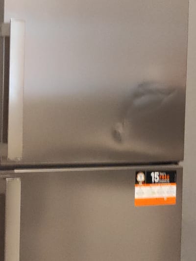 The image shows a close-up of a stainless steel refrigerator door. There is a dent in the upper portion of the door. A sticker with the number "15" and the word "FREE" is visible on the lower portion of the door. The background is out of focus but appears to be an indoor setting. There are no people or signs of activity visible. The location of this image in Chaponost, France is unknown and cannot be determined from the provided visual information.