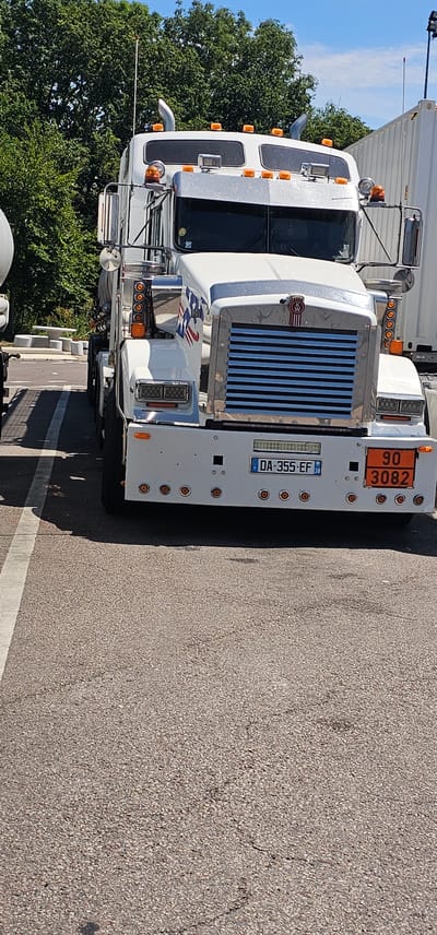 This is a front-on, slightly low-angle shot of a white Kenworth semi-truck parked on asphalt in Vironvay, France. The truck has a chrome grille with blue horizontal slats and the Kenworth logo at the top. It features numerous orange indicator lights along the top of the cab and on the sides. A license plate reading "DA-355-EF" is visible below the grille, with a small circular emblem indicating "38" beneath it. To the right of the license plate is an orange hazard placard displaying "90" at the top and "3082" below. The truck is decorated with a flag decal on the side of the hood, featuring red and blue stripes and white stars. In the background, a white shipping container is visible to the right, and lush green trees form a backdrop to the left and above. The ground is paved with dark asphalt, marked with a white line to the left of the truck. The sky is blue with scattered clouds. No people are present in the image.