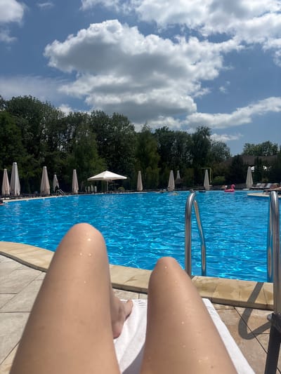 The image shows a first-person perspective of someone relaxing by a swimming pool on a sunny day. The pool is large and filled with bright blue water. Several white umbrellas are set up along the edge of the pool, and lounge chairs are visible behind them. Trees and some buildings form the background. In the foreground, the legs and feet of the person are visible, resting on a white towel. There are small water droplets on their skin, suggesting they have been in the water or recently showered. The sky is a clear blue with fluffy white clouds. The location is identified as Babai, Ukraine.