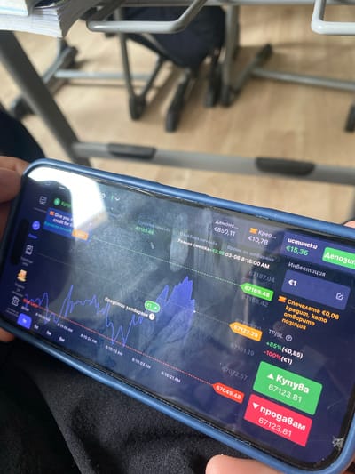 A person's hand holds a dark blue smartphone, displaying a financial trading application in Bulgarian. The phone, encased in a dark blue protective cover, shows a visible crack on the top right of its screen. The app interface features a real-time price chart, options to "Купува" (Buy) and "продавам" (Sell) at 67123.81, and various financial metrics such as "Демонс..." (€850,11), "Кред..." (€10,78), and "истински" (€15,35) balances, likely representing demo, credit, and real accounts. Timestamps like "03-06 8:16:00 AM" and other minute-level data points indicate the activity is occurring in the morning. Labels such as "Инвестиция" (€1), "Очаквана печалба" (Expected profit), "TP/SL" (+85% (€0,85) / -100% (€1)), and "Спечелете €0,06 кредит, като отворите позиция" (Earn €0.06 credit by opening a position) are visible. The background is blurred, showing light-colored flooring and what appears to be metal furniture frames and a stack of papers or a book, suggesting an indoor setting like an office or home in Chirpan, Bulgaria.