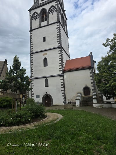 The image depicts the historic St. Nicholas Church (Nikolaikirche) in the city of Meissen, Germany. The church features a prominent white tower with a dark, stepped roof and dark decorative stonework at the corners and around the windows. The tower has several arched windows with decorative tracery. Attached to the tower is a white building with a red tiled roof, which has a dark-shingled spire at the peak. A stone staircase with white balustrades leads up to a dark wooden door on this attached building. To the left of the tower, a portion of another building with a tiled roof is visible. In the foreground, there is a curved pathway made of gravel, bordered by a grassy area with a small tree. Further left, another building with a mansard roof is visible, partially obscured by a leafy green tree. The sky is overcast with scattered clouds. No people or animals are present in the scene. The timestamp "07 липня 2026 р. 2:38 PM" is overlaid on the bottom left corner of the image.