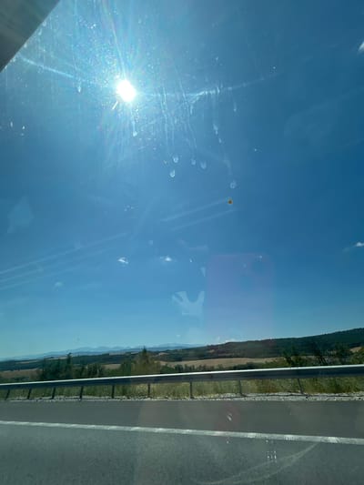 The image depicts a view from inside a vehicle, looking through the windshield on a sunny day. The sky is a clear blue, with the sun prominently visible and casting a bright glare across the upper portion of the image. Streaks and smudges are visible on the windshield, along with some small circular marks. A single, small, yellowish object is also visible in the sky, off-center to the right. In the mid-ground, a highway runs horizontally, bordered by a metal guardrail. Beyond the highway, a rolling landscape unfolds, with fields of dry grass, scattered trees, and distant, hazy mountains. The overall impression is that of a scenic drive through a rural area. The image does not contain any specific details that would place it in the city of Vakarel, Bulgaria.