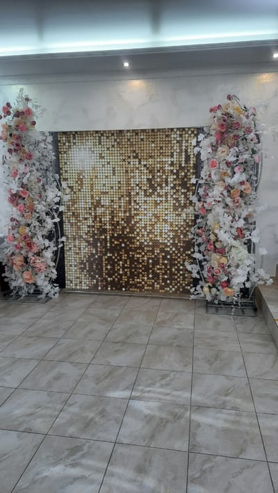 An indoor scene depicts a wedding photo booth setup in what appears to be a banquet hall. The central element is a backdrop composed of gold sequined panels, creating a shimmering, mosaic-like effect. Flanking this backdrop are two large floral arrangements. These arrangements feature a combination of white and light pink roses, along with abundant white foliage and delicate white flowers, cascading down in an arching fashion. The floral structures are supported by dark metal frames.
The floor is tiled with large, light beige tiles that have a marbled pattern. The walls above the sequined backdrop are textured and painted in a light, neutral tone. A decorative cornice runs along the top of the walls, and recessed lighting fixtures are visible in the ceiling.
The overall impression is one of a festive and elegant setting, likely prepared for an event such as a wedding or celebration. The location is identified as Жодзішкаўскі сельскі Савет, Belarus, suggesting this photo booth is situated within a rural council area in Belarus.