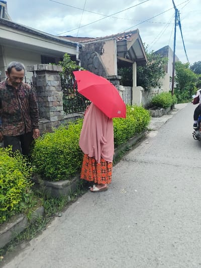 A woman in a pink hijab and orange patterned skirt holds a red umbrella obscuring her face. She stands on a paved street beside a low green hedge and a concrete curb. To her left, a man in a patterned shirt stands by a stone fence. Buildings are visible in the background. A motorbike passes in the distance on the right. The sky is overcast.