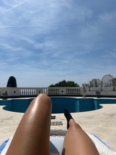 The image captures a person relaxing poolside in Èze, France, under a clear, bright sky. The perspective is from a low angle, looking upwards and across the scene.

In the foreground, the tanned legs of a person are visible, propped up, likely on a lounge chair. One foot, wearing a black low-cut sock, is prominent. The leg closest to the viewer shows a distinct sheen from the sunlight. The person is seated on what appears to be a white wicker chair, partially covered by a blue and white striped towel.

Adjacent to the person is a swimming pool with bright blue water, surrounded by light-colored paving stones. The pool area features an elegant white balustrade with decorative elements, including what look like small urns or planters. Beyond the balustrade, there are lush green trees and architectural features such as white trellised structures, possibly pergolas, and classical-style sculptures or decorative pieces.

The upper portion of the image is dominated by a vast, clear blue sky, showing scattered thin, wispy clouds and several prominent white contrails left by airplanes. The bright, high sun indicates it is midday or early afternoon, contributing to a warm and sunny atmosphere. The overall scene suggests a luxurious and relaxing outdoor setting. No text is visible in the image.