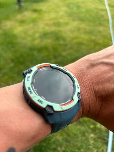 The image shows a close-up of a person's wrist wearing a Garmin smartwatch. The watch has a round, black display surrounded by a light green bezel with orange accents. Several buttons are visible on the bezel, labeled "SET," "BACK," "GPS," "START/STOP," "CTRL," "MENU," and "ABC." The word "GARMIN" is also visible on the watch face. The person's skin is visible, with some fine hairs. The background is out of focus and appears to be a grassy area, with a hint of a white line, possibly a sports field marker. The lighting suggests it is daytime. The image was taken in Haaslava, Estonia.