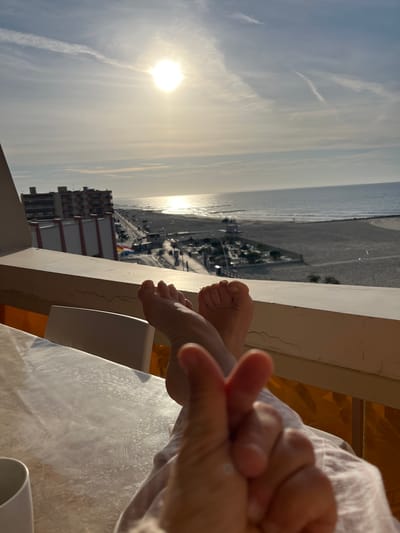 The image shows a view from a high vantage point, possibly a balcony, overlooking Palavas-les-Flots, France. The sea is visible in the background with the sun reflecting on its surface. Buildings, a beach, and a road line the coast. In the foreground, a person is sitting with their feet and hand in view. The person's hand appears to be applying lotion. A white table and part of a chair are also in the frame.
