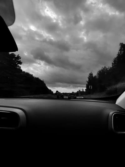 The image captures a view from inside a car, looking forward along a highway. The sky is filled with dramatic, dark clouds, suggesting overcast weather. On either side of the road are tree-lined embankments. Several cars are visible driving ahead, and road signs, including one that appears to indicate a fuel station, are present. The foreground shows the dashboard of the car, including parts of the interior trim and air vents. The location is identified as a highway in Sławków, Poland, though no specific landmarks definitively confirm this without additional context. The scene is entirely in black and white, emphasizing the texture of the clouds and the contrast between the dark car interior and the brighter road and sky.