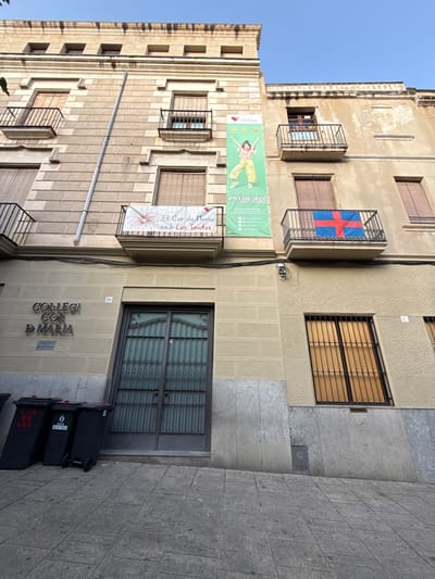 The image shows the exterior of a building in Mataró, Spain, likely a school or religious institution, as indicated by the signage that reads "COL·LEGI COR D'MARIA". The building has a light-colored stone facade with multiple windows and balconies. Two banners are displayed on the building: one reads "EL Cor de Maria amb Les Santes" and features a graphic of fireworks. Another banner, a green one, depicts a young girl in a yellow outfit with the text "APRÈN AMB EMOCIÓ" and social media handles. A flag with a red cross on a blue background is visible on a balcony. In the foreground, three black trash bins are lined up along the sidewalk. The street is paved with square tiles, and the sky above is clear and blue.