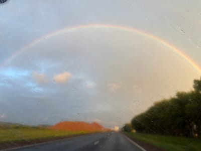 This is a landscape photograph taken from inside a vehicle, looking through a wet windshield. A rainbow arcs across a hazy sky, with clouds visible beneath it. To the left, a vibrant orange mound of dirt or rock is situated beside a green field and a dark asphalt road. On the right side of the road, a dense line of dark green trees stretches into the distance. The overall impression is of a scenic drive through a rural or undeveloped area, possibly following or preceding rainfall. The specific location in Таймурзино, Russia is not depicted or suggested by the visual elements of the image.