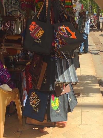 The scene is a street market in Lindi Street, Tanzania (latitude: -3.355, longitude: 37.341). A stack of colorful, handmade tote bags are displayed for sale. The bags feature intricate, patterned fabric designs. A woman wearing a colorful dress is seated in the foreground, while a man in a white shirt and brown pants stands in the background. The street is made of tiled squares, and there is a shadow from a nearby building on the sidewalk. The bags are resting on a wooden table with a brown surface.