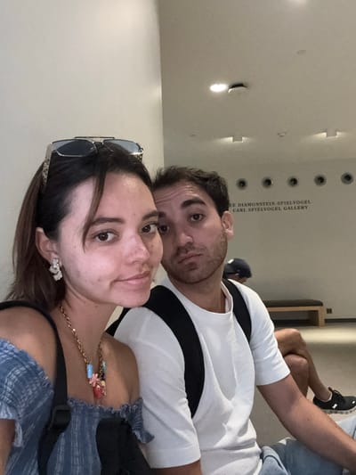 A man and a woman are posing for a selfie in what appears to be a museum or gallery, given the text on the wall behind them which reads "DIAMONSTEIN-SPIELVOGEL CARL SPIELVOGEL GALLERY". The woman is in the foreground, her face close to the camera, and she is wearing a blue off-the-shoulder top, a necklace with colorful charms, and large earrings. She has sunglasses perched on her head. The man is behind her and to her right, also looking towards the camera. He is wearing a white t-shirt and a backpack. In the background, there is a bench and circular lights on the wall. The overall lighting suggests an indoor setting. The location is identified as Washington, United States of America.