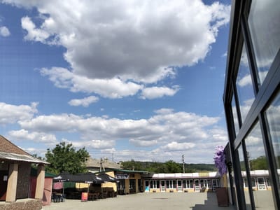 The image captures a daytime view of an outdoor seating area in Congaz, Moldova, likely a cafe or restaurant. The foreground is dominated by the facade of a modern building with large glass windows, reflecting the sky and clouds. To the left, a smaller building with a tiled roof and a covered porch is visible. In the middle ground, several black umbrellas are set up over tables and chairs, suggesting an area for patrons to sit outdoors. Behind the seating area, a row of businesses with white window frames and signs reading "999" are lined up. The background features a green hillside and a blue sky filled with fluffy white and grey clouds. Electrical wires are strung across the scene, indicating an urban or semi-urban setting. No people are visible in the image.