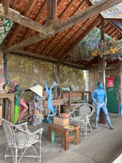 This image depicts an outdoor art studio or exhibition space in Tejakula, Indonesia. The scene is filled with various sculptures, artwork, and furniture. In the foreground, there are several rattan chairs and a wooden table with a small teapot and a yellow object. Dominating the right side of the image is a life-size sculpture of a muscular male figure painted entirely blue, wearing pink briefs, and posed with hands on hips. Behind him, a green door is visible.
Further back, a vibrant green humanoid sculpture stands in a dynamic pose. Next to it, a sculpture of a pink-skinned figure is also displayed. In the center background, a tall, thin blue sculpture is depicted in an upside-down handstand. Various other sculptures and artistic elements are visible on shelves and surfaces, including abstract forms and figurative pieces.
The space is covered by a wooden roof with terracotta tiles, supported by exposed wooden beams. The ceiling is adorned with colorful murals and carved wooden panels depicting human figures and natural scenes. A bamboo blind hangs on the back wall, suggesting an open-air or partially enclosed environment. The floor is made of wooden planks. The overall impression is one of a cluttered but creative artistic space.