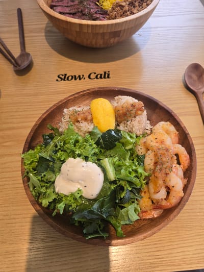 The image shows a wooden bowl filled with a meal on a wooden table. The meal consists of rice, a green salad with dressing, and several pieces of shrimp. A large wooden bowl filled with beef, rice, and a yellow ingredient is visible in the background. The text "Slow, Cali" is visible on the table. This scene is located in the city of Gumi-si, South Korea.