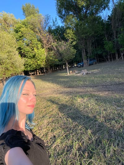 This media file is a selfie taken outdoors, likely during the day. The main subject is a young person with bright blue hair, lying down on the grass. Their face is turned towards the camera, with their eyes closed and a slight smile, suggesting a relaxed or peaceful emotion. They are wearing a black top with ruffled details.

The setting appears to be a grassy area with trees in the background. Sunlight is casting shadows on the grass, indicating it is likely late afternoon or early evening. In the distance, a blue vehicle is partially visible among the trees, suggesting a rural or secluded location. The overall mood is serene and natural. There is no visible text in the image.