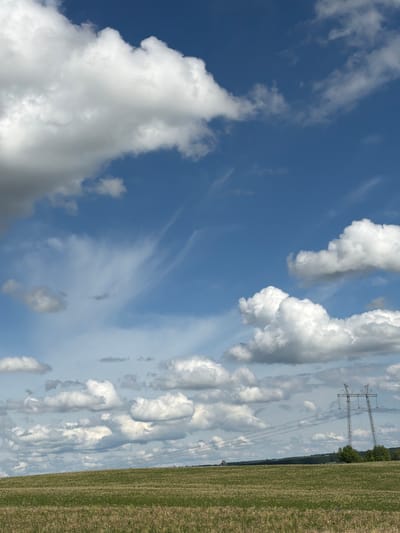The image depicts a rural landscape in Udmurtskoe Kizekovo, Russia. A field of harvested grass occupies the right side of the frame. Power lines and towers stand in the distance, bordering the field. Above, a blue sky is filled with various types of clouds, including cumulus and cirrus formations.