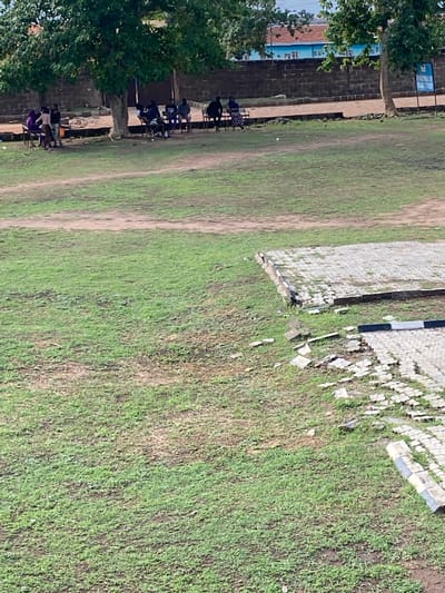 The scene shows a grassy field in Sabo, Nigeria.  A building with blue accents and a red roof adjoins a brick wall along one side of the field.  A row of people sits on chairs against the brick wall, possibly observing or participating in an activity on the field.  The opposite side of the field features a stone structure, partially in disrepair, with scattered stones and debris in the adjacent grass. The field is predominantly green with visible paths or worn areas.  Trees are present near the building.