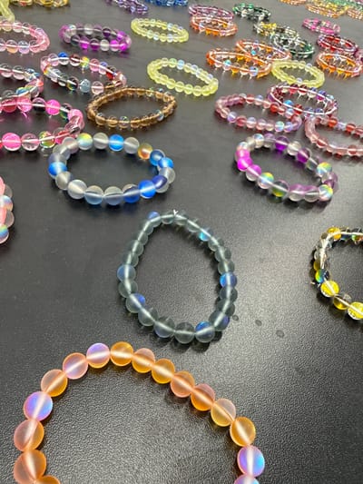 The image shows a collection of beaded bracelets on a black surface. The bracelets are of various colors and designs, including translucent beads with iridescent reflections, beads with internal wire structures, and solid colored beads. The bracelets are arranged somewhat randomly on the surface. There are no people visible in the image. The location of the image is unknown and not obviously deducible from the visual information. The supposed location of Синьково, Russia is not apparent within this image.
