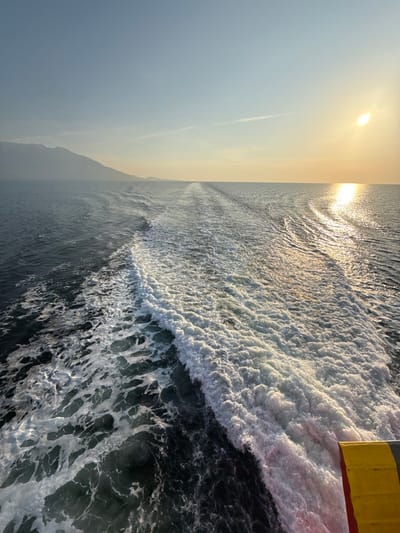 The image displays an expansive maritime scene viewed from the stern of a moving vessel, likely a ferry, consistent with the coastal region near Kato Karioutes, Greece. The primary subject is the turbulent white wake generated by the vessel, which dominates the midground and extends towards the horizon. The water on the left side of the wake is dark blue-green, while the water on the right reflects a bright golden light from the sun. The sun, a distinct golden orb, is positioned low on the right horizon, casting a shimmering path across the water. Along the left horizon, a long, dark, and hazy mountainous landmass is visible. The sky transitions from a pale blue in the upper left to a yellowish-orange near the sun, with a faint linear contrail present in the upper right. A segment of the vessel's stern, featuring yellow and red-orange colors, is partially visible in the bottom right corner of the frame. No individuals, other vessels, or discernible actions beyond the vessel's movement are present.