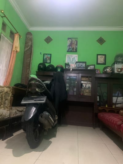 The image shows the interior of a room in a house located in Lagadar, Indonesia (latitude: -6.928, longitude: 107.535). The room has green walls with white trim, and a white ceiling. There are numerous framed photographs and pictures hanging on the walls. A brown wooden cabinet is positioned against the wall to the right of the room, and it is filled with various items, including books, and other objects. Three helmets, two black and one green, are sitting on top of the cabinet. An electric rice cooker with the brand "MAGIC" is visible on the cabinet’s top shelf. A black motorcycle is parked next to the cabinet, facing away from the camera. It appears to be a Yamaha motorcycle. A red couch sits in the foreground to the right of the room, with a small table in front of it. The motorcycle is blocking the majority of the view of the room. The image was taken from the perspective of a person standing in the center of the room, looking towards the back wall. The light source appears to be artificial, and the image is slightly blurry.