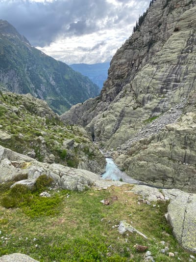 The image displays a mountainous landscape within a deep valley, characteristic of the Swiss Alps near Innertkirchen. The scene is dominated by steep rock formations and a flowing river under an overcast sky.

The sky occupies the upper portion, featuring heavy, dark grey clouds interspersed with areas of lighter grey and white, suggesting broken cloud cover or an impending weather front.

To the left, a large mountain slope rises steeply, covered predominantly with dense green vegetation, including trees and grass, with visible rock outcrops and some lighter patches near the summit. In the distant background, between the two main mountain flanks, additional hazy blue mountain ranges are visible.

To the right, a massive, imposing rock face dominates, appearing closer than the left slope. This cliff is characterized by layered, light grey-green rock with vertical dark streaks, possibly from water erosion or mineral deposits. Small patches of evergreen trees cling to the upper sections and ledges. A substantial scree slope, composed of loose grey rocks and debris, extends from the mid-section of this cliff down towards the valley floor.

The center of the image opens into a narrow gorge where a river flows. The river exhibits a distinct light turquoise or glacial blue color, indicating its origin from glacial meltwater. Its bed is composed of light-colored rocks and gravel.

The immediate foreground is a rough, uneven terrain consisting of large, irregular boulders covered in moss and lichen, interspersed with patches of vibrant green grass and low-lying shrubs. In the lower right corner, a small, dark brown, irregularly shaped object rests on the grass. A small cairn or pile of white stones is also present in the grassy area slightly left of the bottom center.

No human figures or distinct man-made structures are visible. The environment is rugged and natural, reflecting an alpine setting.