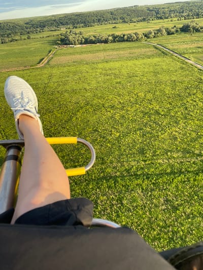 An aerial perspective captures a person's legs and torso from the perspective of a hot air balloon basket, with the vast green landscape of Turovo, Russia stretching out below. The subject is wearing white sneakers and black shorts, with their right leg extended and resting on a yellow-padded handle. The balloon basket itself is partially visible, appearing to be constructed from metal and fabric. The expansive countryside below consists of lush green fields, dotted with patches of trees and agricultural land. A dirt road winds through the fields, leading towards a distant treeline that borders the horizon under a pale, cloudy sky.