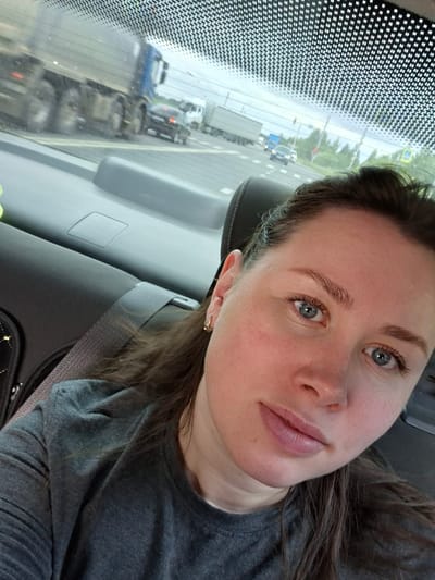 A woman is taking a selfie from the backseat of a vehicle. The woman has light skin, blue eyes, and brown hair. She is wearing a gray t-shirt and a gold earring. The interior of the car is visible, including a headrest, a seatbelt, and the car's interior paneling. Through the rear window, a highway scene is visible with several trucks and cars driving on the road. The sky is overcast. The location is identified as Novaya Kazminka, Russia.