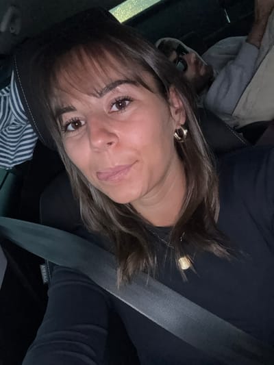 This image captures the interior of a vehicle in Reventin-Vaugris, France. In the foreground, a female subject with medium-length brown hair and bangs looks directly at the camera with a slight smile, her tongue partially protruding. She wears a dark long-sleeved top, gold hoop earrings, and a gold pendant necklace. A grey seatbelt, labeled "AIRBAG," diagonally crosses her chest. To the upper right, a male subject is partially visible in the background, leaning against a seat, wearing a light-colored hooded top and dark sunglasses. A striped garment is visible to the left of the primary subject. The lighting indicates a low-light environment, possibly nighttime.