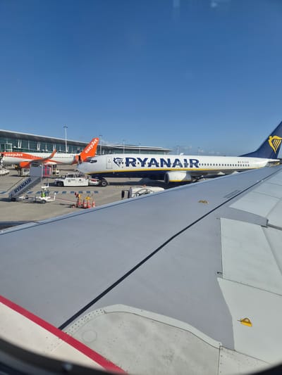 The image shows a view from an airplane window. The wing of the plane occupies the right side of the frame, with a visible red and white stripe along the edge. Beyond the wing, on the tarmac, there is another airplane painted in the colors of the airline "Ryanair" and an "EasyJet" plane behind it, along with ground support equipment, including stairs and vehicles. The terminal building is visible in the background. The sky is clear and blue. The location is in Moreira, Portugal, identified by the presence of Ryanair and EasyJet airlines, which are frequent at Portuguese airports.