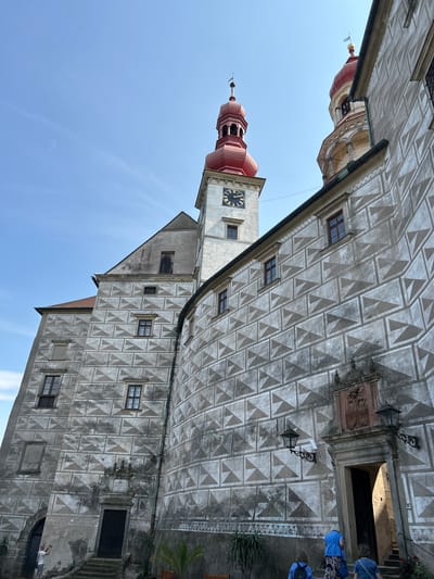 The image shows a view of Náchod Castle in Náchod, Czechia. The castle features striking sgraffito decoration on its exterior walls, characterized by a repeating geometric pattern of triangles in shades of gray and white. Two distinct towers are visible: one with a clock face and a red onion dome, and another with a similar red dome. Several people are present in the scene. In the foreground, three individuals, two wearing blue shirts and one in a floral skirt, ascend a stone staircase towards an arched doorway. To the left, a man in a white t-shirt and shorts stands near an entrance, holding a camera. The sky above is clear blue with a few faint clouds. The architecture suggests a historical and well-preserved structure.
