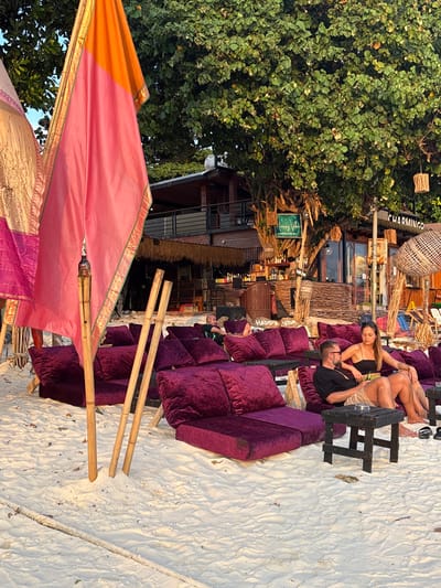 The image captures a beachside leisure area in Ban Ko Li Pe, Thailand, during golden hour. The foreground features fine white sand, marked with subtle undulations and footprints. On the left, tall bamboo poles support colorful fabric banners in shades of pink, orange, and gold. In the midground, multiple dark purple velvet or velour lounge chairs and ottomans are arranged directly on the sand.
A male and a female are seated on one of these lounge arrangements on the right. The male wears a black t-shirt, tan shorts, dark-rimmed glasses, and has a beard; he is looking down at a small rectangular object in his hands while holding a green bottle. The female, seated next to him, wears a black strapless top and has long dark hair, looking in his direction. Both are barefoot, with black sandals visible near the female's feet. A small, dark wooden square table stands in front of them. To their left, a second male is visible, lying back on a purple lounge with his feet elevated.
The background reveals a two-story establishment with a dark wooden and brick facade, partially covered by a thatched roof. A large, densely leafed green tree dominates the center background, with various woven decorations or lamps hanging from its branches and the building structure. Signs visible on or near the building include "CHARMINGO" and "HAPPY VIBE," suggesting a bar or restaurant setting with bottles lining a counter. The overall scene depicts a relaxed environment at a beachfront venue.