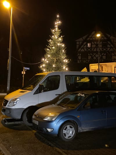 A nighttime scene features a brightly lit Christmas tree next to a half-timbered building. A blue car and a white van are parked beside the tree. A streetlamp illuminates the scene from the left. Street signs are also visible near the base of the tree.