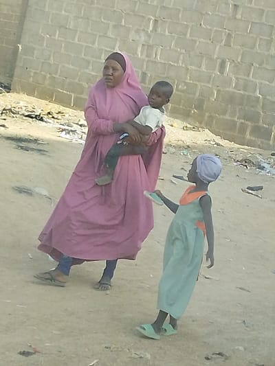 A woman in a long, dusty-rose robe and headscarf carries a small child in a white shirt. Both are dark-skinned. A younger child wearing a light green dress, light green headscarf, and matching sandals stands in front of them holding a sandal in their left hand. All three are on a dirt road. A brick wall is in the background, along with some scattered litter.