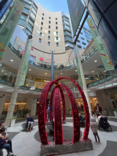 The image depicts the multi-story atrium of the Bay Street Complex in Saint Julian's, Malta, viewed from a low angle looking upwards. The central focus is a large, freestanding festive decoration in the foreground, composed of several interconnected, arch-shaped structures covered in deep red, glittery material and illuminated with numerous small red lights. The base of this decoration is adorned with silver tinsel.
The atrium features multiple levels of commercial establishments, with glass railings and shopfronts visible on each floor. Signage for "Pizza Hut" and "L'Occitane" is discernible on upper levels, while "KARL LAGERFELD," "MAX & CO.," "TED BAKER," and "SKIPPER" are visible on lower levels. String lights are draped along the railings, suggesting a decorative theme. Tall vertical banners displaying "Your daily oasis" text are attached to pillars. The upper levels of the complex feature a light-colored, curved facade with rows of rectangular windows, characteristic of a hotel or office building. Natural light enters from an open-air or glass-roofed section above.
Several individuals are present in the foreground and midground. To the left, a woman is seated on a bench next to a child, who is also seated. Another child in a blue jacket is seated on a black, curved bench structure. To the right, a child in a light-colored hooded jacket stands with their back to the camera, facing the red arch decoration. Behind them, an adult pushes a dark-colored baby stroller containing an infant. Another person is partially visible seated on a black curved bench behind the red arches. The floor is tiled in geometric patterns of light and dark grey.