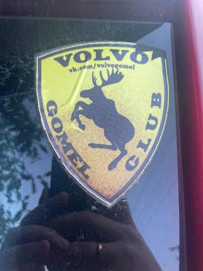 A yellow shield-shaped sticker is adhered to the back window of a dark-colored vehicle. The sticker features a silhouette of a moose, with the text "VOLVO" in large capital letters above it. Below "VOLVO" is the web address "vk.com/volvogomel". The words "GOMEL" and "CLUB" are curved along the left and right edges of the shield, respectively. The sticker appears weathered and slightly peeling at the edges. The overall scene is dimly lit, with reflections on the glass indicating an outdoor environment. The vehicle is likely parked on a street or in a lot in Бабовіцкі сельскі Савет, Belarus, given the text on the sticker.