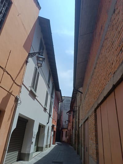 The image depicts a narrow street in Casalmaggiore, Italy, flanked by buildings on both sides. The street is paved with cobblestones and illuminated by sunlight casting shadows.

On the left side of the street, an orange building is visible, followed by a white building with shutters and an ornate lamppost. Below the windows of the white building, there are roll-up doors and arched doorways. The building on the right is constructed of red brick and features large wooden doors.

The sky is a clear blue with scattered clouds. The perspective of the image suggests a low camera angle, emphasizing the height of the buildings and the narrowness of the alleyway. No people are present in the scene. The overall impression is that of a quiet, historic street in an Italian town.
