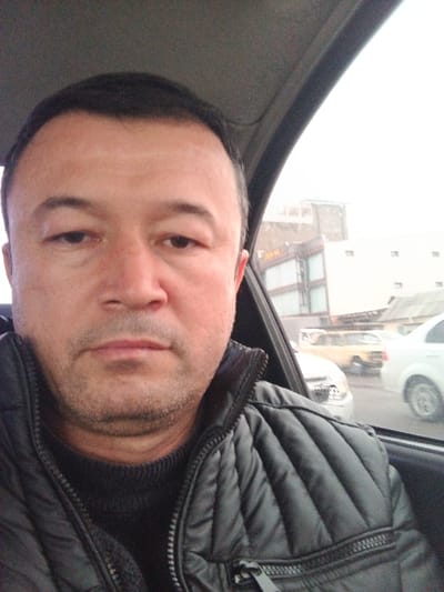 A middle-aged man with dark hair, brown eyes, and tan skin sits inside a car, wearing a black quilted jacket over a black sweater. He is looking at the camera. Outside the car window, a building with multiple stories and a dark sign is visible. Several cars, including a white sedan, are also seen outside. The weather appears overcast.
