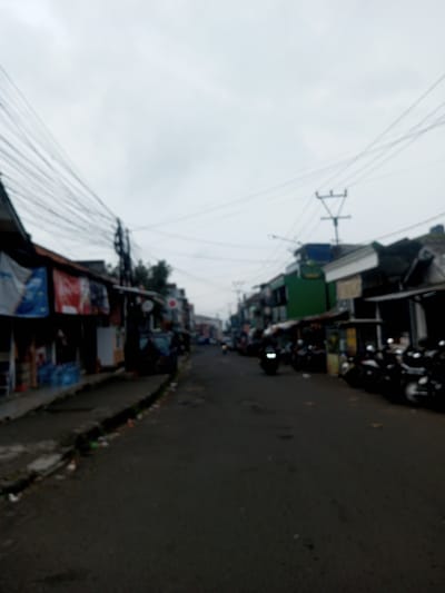 The image displays an indistinct, overcast street scene in Houaythat, Laos, observed from a low perspective looking down a paved thoroughfare. The asphalt road, appearing dark grey, recedes into the background. A concrete curb and narrow dirt strip line the left side of the road, showing scattered small debris. On both sides of the street, rows of two-story structures are visible. The buildings on the left feature multiple awnings and various banners, suggesting commercial establishments. Overhead, a dense network of dark electrical wires spans the street, attached to utility poles. On the right side of the road, a significant number of motorcycles are parked parallel to the curb. Further down the street on the right, a single motorcycle with an illuminated headlight is in motion, approaching the viewer. The sky is uniformly light grey, indicating an overcast day. No distinct individuals or specific human actions are discernible due to the image's overall blurriness.