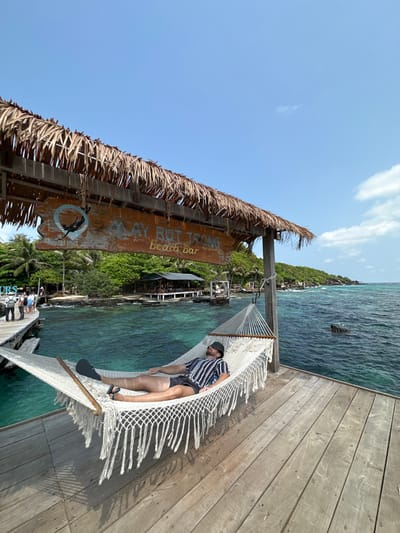 The image captures an outdoor scene at the Mây Rút Trong beach bar, situated on Mây Rút Trong Island in the An Thới Archipelago, near Thành phố Phú Quốc, Vietnam, under a clear blue sky with minimal cloud cover. A Caucasian male, wearing a black and white striped short-sleeve shirt, dark shorts, and dark flip-flops, rests horizontally in a white woven hammock with tassels. A dark bucket hat covers his eyes. The hammock is suspended between two wooden posts on a weathered wooden pier. Above the hammock, a thatched roof shelters a rustic wooden sign that reads "MÂY RÚT TRONG beach bar" and features a circular logo depicting a person in water. In the background, clear turquoise ocean water transitions to darker blue further out. A wooden walkway extends into the water to the left, where several indistinct figures (people) are visible. Beyond the water, a building with a dark roof and open sides is visible on stilts, surrounded by dense green tropical vegetation along a rocky shoreline. Submerged rocks are discernible in the deeper water.