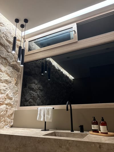 The image shows a bathroom sink in a modern, minimalist design. The sink is made of light brown stone and sits on a countertop of the same material. There is a large window with a frosted glass pane and a mirror above the sink. Three black light fixtures are hanging from the ceiling and are reflected in the mirror. The mirror reflects a stone wall behind the bathroom. There are two bottles of hand soap on a wooden tray next to the sink, and a white towel is hanging on a rack above the sink. The faucet is black, and there is a black drain stopper on the counter. The scene appears to be in a bathroom in a high-end, modern hotel or residence in Santa Fe, Costa Rica, at the coordinates 9.647 latitude and -85.166 longitude.